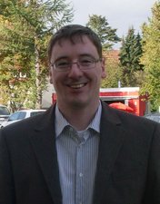 Freundliches Porträt eines Mannes im Anzug Ein lächelnder Mann mit Brille trägt einen Anzug und steht vor einer grünen Landschaft mit Bäumen und Autos im Hintergrund.