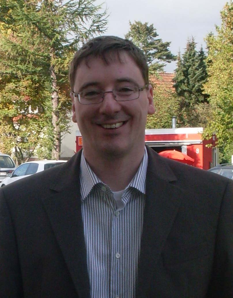 Ein lächelnder Mann mit Brille trägt einen Anzug und steht vor einer grünen Landschaft mit Bäumen und Autos im Hintergrund.