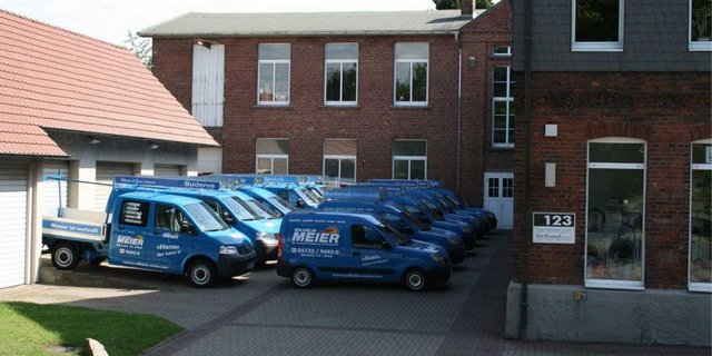 Eine Reihe blauer Lieferwagen parkt vor einem Backsteingebäude mit großen Fenstern und einem grauen Dach.